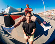 © Studio Marmellata - A fisheye view of two men in an outdoor urban environment, with one man seated on a ledge and stretching his legs over the other standing man below, under a clear blue sky