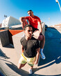 © Studio Marmellata - Two young men, one sitting on a ledge in a red shirt and the other in yellow shorts, in an outdoor urban space under a clear blue sky with a fisheye perspective capturing the setting.