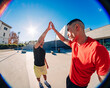 © Studio Marmellata - Two young men, one in a red shirt and one in black, exchanging a high-five outdoors with a fisheye lens capturing the vibrant sunny day and urban setting in the background.