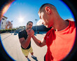 © Studio Marmellata - Two young men doing sport activity captured in a fisheye lens perspective, one wearing a red shirt and the other in black, engaging in a handshake under a sunny blue sky