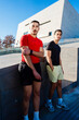 © Studio Marmellata - Two male runners one in red and one in black, pose confidently near a modern architectural building under a clear blue sky on a sunny day, exuding style and camaraderie.
