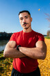 © Studio Marmellata - Male Runner wearing a bright red shirt stands confidently with arms crossed on a sunny autumn day, surrounded by golden fallen leaves and a calm river in the background.