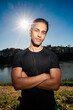 © Studio Marmellata - man doing sport in a black shirt standing confidently with arms crossed as the sun creates a starburst effect behind him in a scenic outdoor setting near a calm river under a bright blue sky.