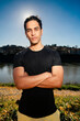 © Studio Marmellata - man doing sport in a black shirt standing confidently with arms crossed as the sun creates a starburst effect behind him in a scenic outdoor setting near a calm river under a bright blue sky.