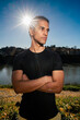© Studio Marmellata - man doing sport in a black shirt standing confidently with arms crossed as the sun creates a starburst effect behind him in a scenic outdoor setting near a calm river under a bright blue sky.