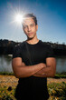 © Studio Marmellata - man doing sport in a black shirt standing confidently with arms crossed as the sun creates a starburst effect behind him in a scenic outdoor setting near a calm river under a bright blue sky.