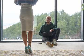 A blonde man sitting casually on the floor of a modern office, leaning against a window as he uses his laptop, with his attractive female colleague standing nearby