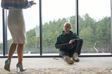 A blonde man sitting casually on the floor of a modern office, leaning against a window as he uses his laptop, with his attractive female colleague standing nearby