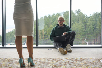 A blonde man sitting casually on the floor of a modern office, leaning against a window as he uses his laptop, with his attractive female colleague standing nearby