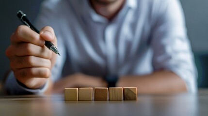 Fototapete - A person holds a pen, poised to write, while focusing on wooden blocks arranged on a table, suggesting creativity or planning.