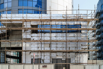 Wall Mural - A modern city building under construction, covered in scaffolding. The building's facade is partially covered in white insulation panels. Scaffolding on Modern Building Facade. Construction Site.