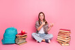 © deagreez - Young girl in casual outfit sitting with books and backpack on pink background, smiling and pointing