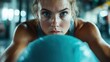 © Dulemegapixel - A dedicated female athlete is intensely focused, performing an exercise with a fitness ball, showcasing strength and determination in a gym environment.
