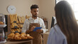 © Krakenimages.com - Man in brown apron using tablet with woman customer in bakery shop interior displaying various bread and pastries on shelves against the clock wall at daytime