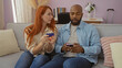 © Krakenimages.com - Interracial couple sitting together on living room couch with woman holding credit card and man using smartphone in cozy home setting