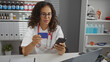 © Krakenimages.com - Woman in clinic holding credit card and smartphone wears glasses and stethoscope surrounded by medical supplies and white uniform focused on online transaction work