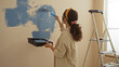 © Krakenimages.com - Woman painting wall indoors, using roller to apply blue paint, homemade renovation project in living room, showcasing creative home improvement efforts.