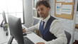 © Krakenimages.com - Young man in office setting, wearing glasses and vest, sits focused at a desk with computer and documents, showcasing a modern and professional workplace environment.