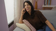 © Krakenimages.com - Woman talking on phone in cozy living room with brown shirt sitting on gray sofa next to large window with books on shelf in background