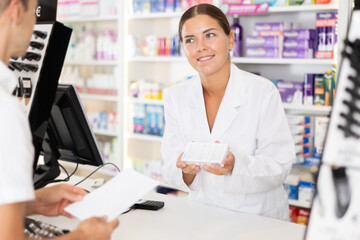 Wall Mural - Affable young woman chemist helping buyer with prescribed medical preparation in pharmacy store