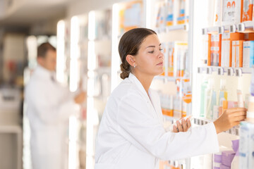 Wall Mural - Positive young female pharmacist placing medical product on shelf in chemist's shop