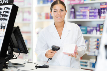 Girl pharmacist in cash area of pharmacy works with computer and goods, scans drugs, checks availability. Chemists shop has wide selection of cosmetics, care products, medicines