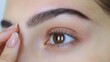 © MetaPixel - Close up of female hand showcasing microblading technique on eyebrow with client observing prior to permanent tattoo application and design