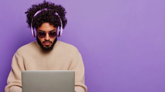 Curly haired man wearing sunglasses and headphones using laptop on violet background with ample copyspace for text casual entertainment theme