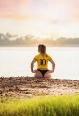 Wall Mural - brazilian woman in a yellow jersey sits by a serene lakeside at sunset, enjoying the tranquil view.
