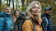 © The 2R Artificiality - Blonde middle-aged woman in golden jacket with backpack smiles confidently while hiking with diverse friends in lush green woods, ample copyspace for text.