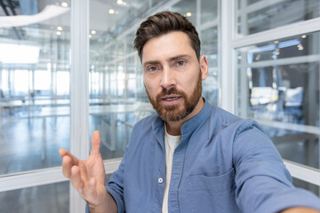 Poster - Man using video calling app on smartphone, office worker looking into camera and talking to colleagues and partners remotely while sitting at desk inside office.