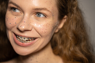 Funny young woman covered with glitter, licks metal braces, beauty portrait close-up