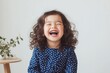 © World of AI - six year old girl with medium brown skin and curly pigtails, wearing a blue polka-dot dress, sitting on a stool and laughing, set against a pastel pink background.