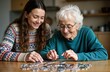© Pete - Young caregiver happily assists senior woman putting puzzle pieces together at home. Senior woman smiles contentedly during activity. Caregiver helps with puzzle. Domestic scene of care, leisure.