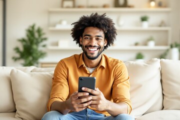 Wall Mural - Smiling man holding smartphone while sitting on a sofa in cozy living room with blurred background.