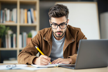 Poster - Young man studying and writing notes at a desk with a laptop.