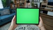 © Adri - Hands holding tablet with green screen in living room.