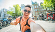 © Davide Angelini - Happy tourist taking selfie picture in Amsterdam, Netherlands - Cheerful man using smart mobile phone device outside - Student traveler enjoying summer european vacation - Life style tourism concept