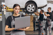 © anatoliycherkas - Worker of autoservice, beautiful young woman holding laptop in hands and looking at camera, smiling. Mechanics fixing, repairing automobile lifted on bridge behind.