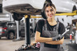 © anatoliycherkas - Smiling young woman mechanic standing beneath lifted car, holding tools and looking at camera in car service garage
