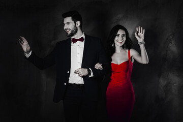 Poster - Portrait of hollywood lady in tight violet dress, handsome vip man in tuxedo with bow attractive superstars holding hands go along the red carpet wagging with palm isolated grey background spotlight