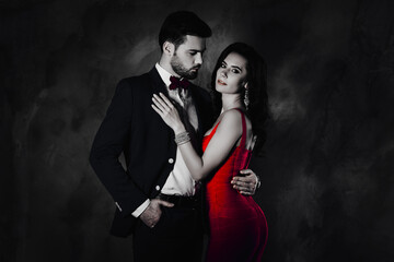 Poster - Portrait of man with stubble in tux with bowtie holding hand in pocket of pants looking at his charming, pretty lover with spouses hugging over dark grey background. Intimacy vogue glamour concept