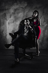 Poster - Portrait of rich gentleman in formal wear sitting in leather chair holding glass with cognac and cigar in hands, pretty charming lady in purple dress standing near isolated on grey background