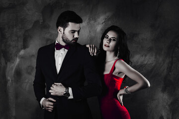 Poster - Portrait of virile man with stubble in tuxedo with bowtie correcting fixing buttons on his jacket looking at his charming tempting rich wealthy lover with hairstyle curls isolated on grey background