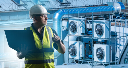 Man engineer servicing conditioning equipment. Ventilation units. Man with laptop controls conditioners. Ventilation equipment on roof building. Worker in yellow vest adjusts air conditioners