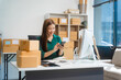 © Phushutter - An Asian young woman sits in a room, working on a computer and making calls or video calls, managing her small business and online sales, surrounded by cardboard boxes ready for shipping.