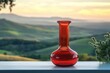 © Vadym - Red glass vase sits on a white table against a backdrop of rolling hills at sunset. Scenic rural landscape complements the elegant object. Rural tranquility and tranquil outdoor scenery.