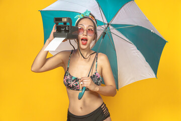 Wall Mural - Emotional Asian girl in swimsuit with headband under umbrella with instant camera. Isolated