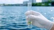© Moopingz - A hand in a glove holds a test tube with a green sample, against a backdrop of water and buildings, suggesting environmental testing or research.