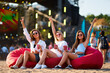 © artiemedvedev - Girls sit on red bean bag chairs at beach concert, raising drinks in toast, blue sky. Enjoy summer music event, audience in background, casual style, sunglasses, joyful vibe, friends fun time.